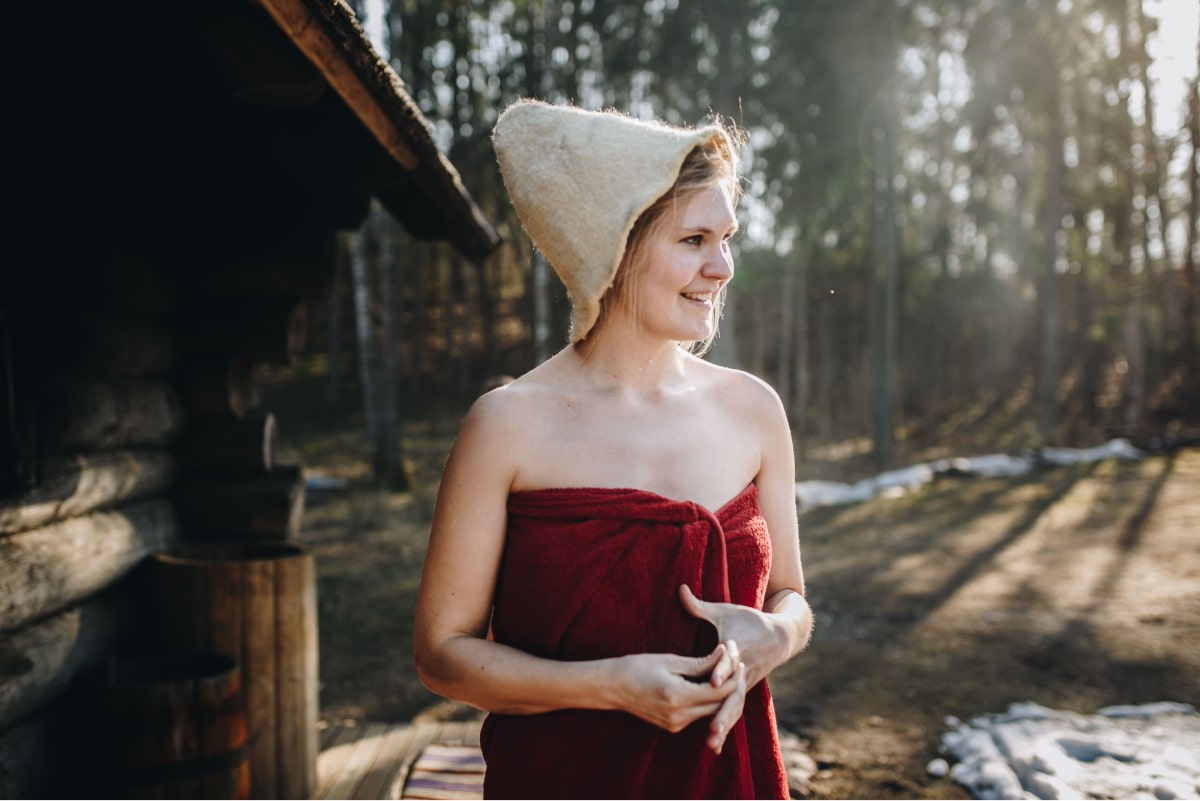A woman wearing a traditional felt sauna hat outside a log sauna cabin